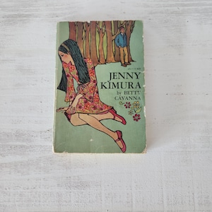 May include: Vintage paperback book titled "Jenny Kimura" by Betty Cavanna. The cover features an illustration of a woman in a floral dress and red shoes, with a boy and trees in the background. The book has a green cover.