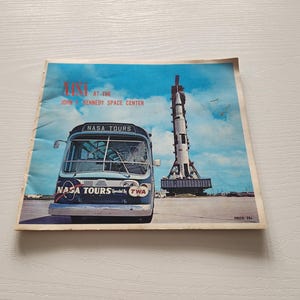 May include: Vintage NASA Tours brochure featuring a blue and white bus with "NASA TOURS" printed on the front. The brochure shows a rocket on a launch pad against a blue sky. The text "NASA at the John F. Kennedy Space Center" is visible.