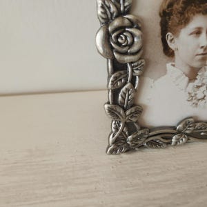 May include: A vintage silver-toned picture frame with a rose and leaf design. The frame holds a sepia-toned photograph of a woman. The frame is on a white surface.