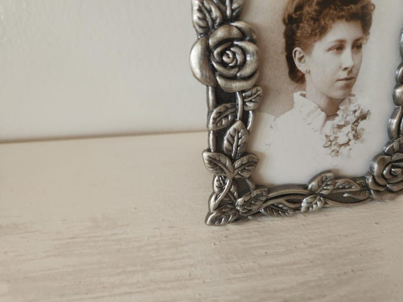 May include: A vintage silver-toned picture frame with a rose and leaf design. The frame holds a sepia-toned photograph of a woman. The frame is on a white surface.
