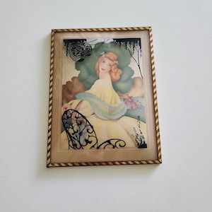 May include: Framed vintage art print featuring a woman in a yellow dress, with red hair, and a blue shawl. The artwork has a decorative frame with a brown and gold striped pattern. The background includes a green tree and a small figure.