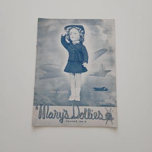 May include: Vintage blue-toned cover of "Mary's Dollies" Volume No. 6. The cover features an illustration of a doll in a navy blue outfit, saluting, with airplanes in the background. The text "The Wavette" is above the doll.
