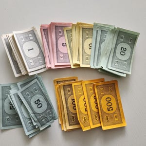 May include: Stacks of Monopoly money in various denominations, including $1, $5, $10, $20, $50, and $500 bills. The bills are in different colors, such as white, pink, yellow, green, and gold. A classic board game accessory.