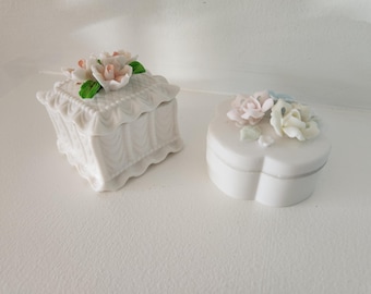 May include: Two white porcelain trinket boxes with floral designs. One box is square with a floral design on the lid. The other box is heart-shaped with a floral design on the front.