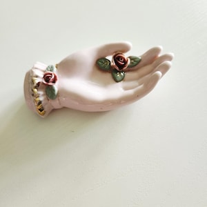 May include: A pink ceramic hand-shaped dish with two red rose accents and green leaves. The wrist area has gold trim. This decorative piece is likely used for holding small items.