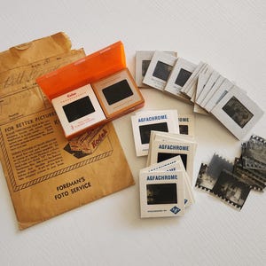 May include: A collection of vintage photography slides and film. Includes an orange Kodak slide case, several Agfachrome slide mounts, and a brown paper envelope with text that reads "Foreman's Foto Service."