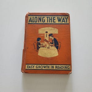 May include: An antique book titled "Along the Way" with the subtitle "Easy Growth in Reading." The cover is orange with black and gold lettering and an illustration of two people at a table.