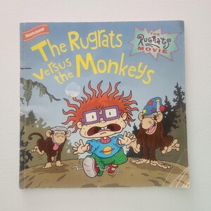 May include: A book cover featuring the Nickelodeon cartoon characters, the Rugrats, in a jungle setting. The title of the book is "The Rugrats Versus the Monkeys" and the subtitle is "The Rugrats Movie". The cover features Tommy Pickles, Chuckie Finster, and a monkey.