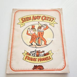 May include: Vintage children's book titled "Seen Any Cats?" featuring illustrations of two figures in orange outfits and a group of cats. The cover has a cream background with orange and yellow accents and the author's name, Frank Modell.