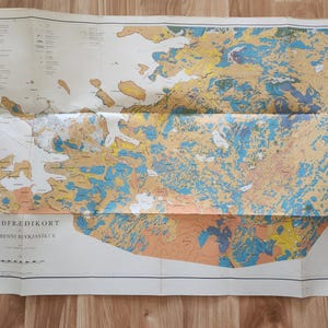 May include: Folded vintage map of a geographical area, featuring a detailed illustration with a variety of colors including blue, yellow, and orange. The map has text in a foreign language and is displayed on a wooden surface.