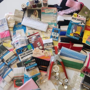 May include: A collection of sewing notions including various spools of thread, packages of Wrights seam binding, quilt binding, hem facing, iron-on sock patches, Dills pipe cleaners, elastic cord, eyelets, picture hangers, and a metal hole punch tool.