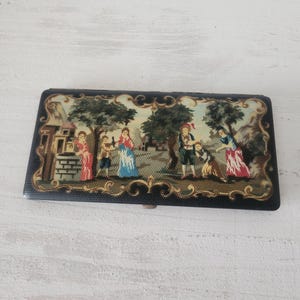 May include: A rectangular, black case with a detailed tapestry scene of people in a park. The tapestry features figures in period clothing, trees, and a building. The case has a decorative gold-colored border.