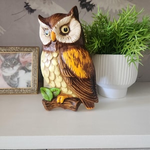 May include: A ceramic owl figurine with brown, yellow, and cream coloring. The owl is perched on a brown branch with green leaves. The owl has large yellow eyes and is a decorative item.