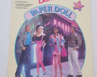 May include: A Barbie Paper Doll book with the title "Barbie Paper Doll" in a pink and blue design. The cover features illustrations of Barbie dolls and the text "Includes 4 Paper Dolls", "4 Paper Dolls", "15 Precut Fashions and Accessories", and "Fashion Tote".