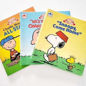 May include: Three paperback children's books featuring the Peanuts characters Snoopy and Charlie Brown. The books are titled "Charlie Brown All-Star", "He's Your Charlie Brown", and "Snoopy, Come Home".