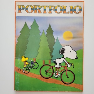 May include: Vintage "Portfolio" book cover featuring Snoopy and Woodstock riding bicycles in a forest scene. The cover has the word "PORTFOLIO" in large, colorful letters at the top.