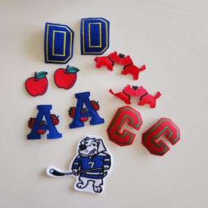 May include: Assortment of embroidered patches featuring letters, apples, and a hockey-playing dog. The letters include two blue "D"s with gold trim, two red "C"s with green trim, and two blue "A"s with red tomatoes.