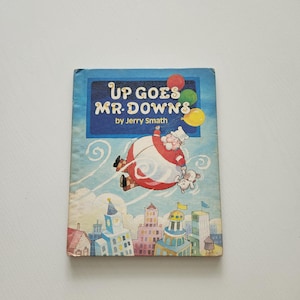 May include: A hardcover children's book titled Up Goes Mr. Downs by Jerry Smath. The cover features a blue sky background with a man floating while holding a small dog, above a colorful city skyline. Red, green, and yellow balloons are visible.