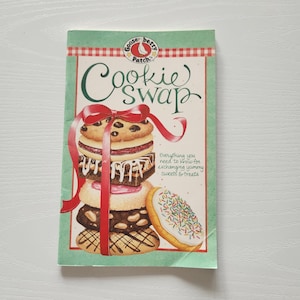 May include: A cookbook titled "Cookie Swap" with a stack of cookies on the cover, tied with a red ribbon. The cover features the text "Everything you need to know for exchanging yummy sweets & treats."