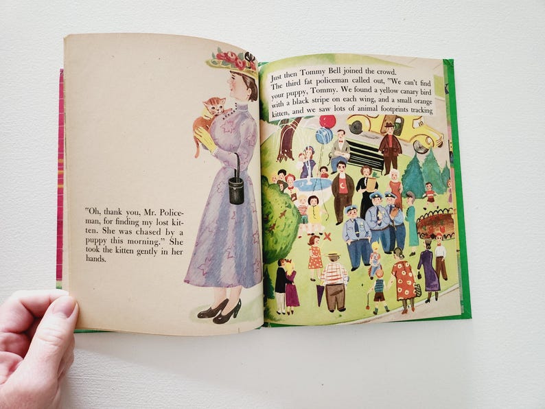 Może przedstawiać: Antyczna książka dla dzieci otwarta na stronie z ilustracją kobiety trzymającej kociaka. Tekst na stronie brzmi: "Och, dziękuję, panie policjancie, za odnalezienie mojego zaginionego kociaka. Był ścigany przez szczeniaka tego ranka." Ona wzięła kociaka delikatnie w swoje ręce. Przeciwległa strona przedstawia ilustrację tłumu ludzi, w tym policjant&oacute;w, dzieci i zwierząt.