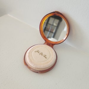 May include: A vintage compact with a brown plastic case and a mirror. The compact has a white puff and the words "John Robert Powers" written in gold lettering.