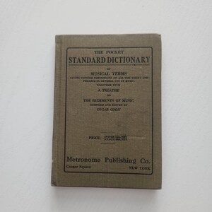 May include: A vintage pocket-sized book titled "The Pocket Standard Dictionary of Musical Terms." The cover is a muted olive green with black text. The book includes a treatise on the rudiments of music, compiled and edited by Oscar Coon.