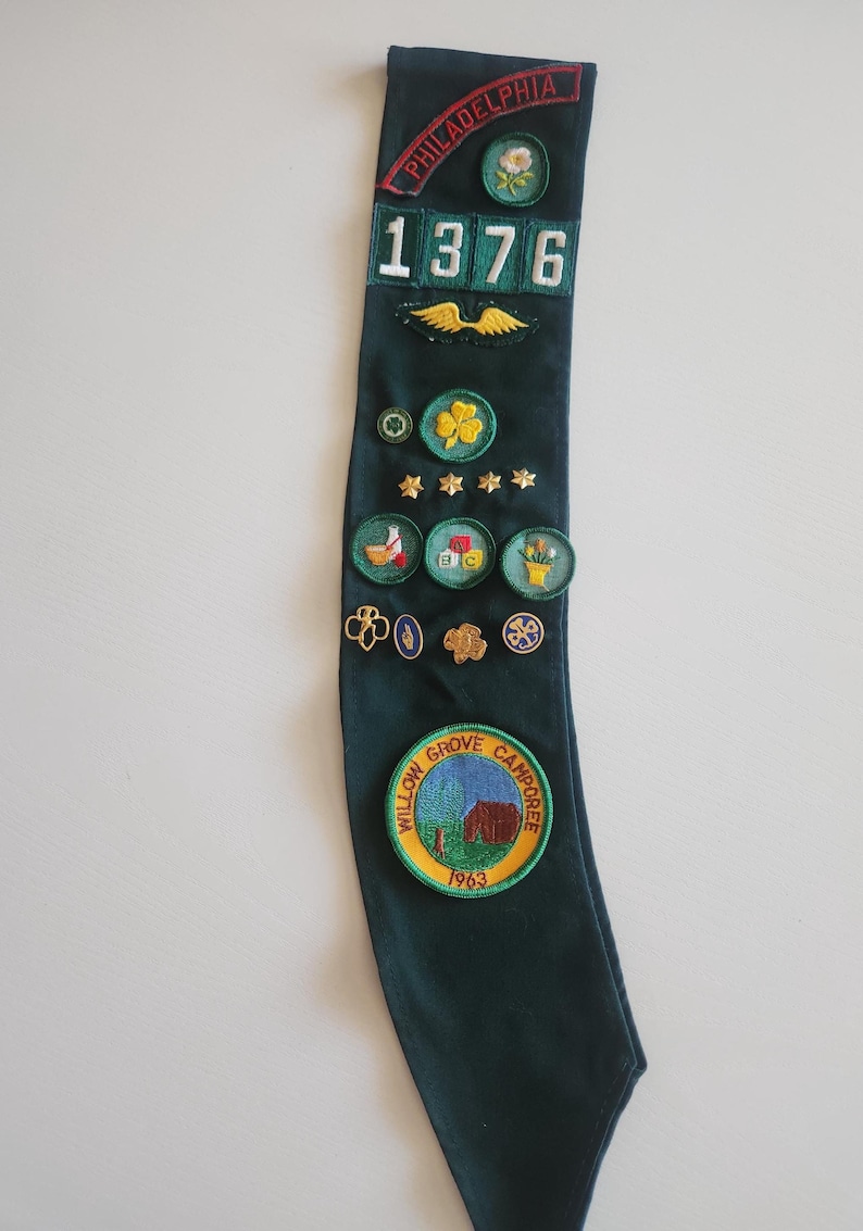 May include: A dark green sash with various embroidered patches and pins. Patches include the word "PHILADELPHIA" and the number "1376". Other patches feature a clover, stars, and a camp scene. A vintage collectible.