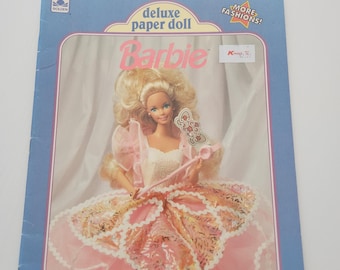 May include: A vintage Barbie deluxe paper doll set. The cover shows a Barbie doll in a pink gown with a wand. The packaging is blue with the words "deluxe paper doll Barbie" and "MORE FASHIONS!" printed on it. The Golden logo is in the upper left corner.
