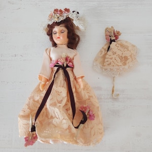 May include: Two vintage dolls. The larger doll wears a peach-colored dress with lace overlay, a dark brown ribbon, and floral accents. The doll has brown hair and a floral hat. The smaller doll has a similar dress and floral accents.