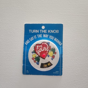 May include: A vintage button with the text "I'm yours" inside a red heart, surrounded by colorful hearts. The button is on a blue card that reads "Turn the knob and say it the way you wanna".