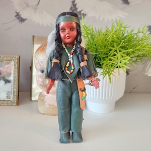 May include: Vintage doll with a Native American theme. The doll has a dark complexion, black braided hair, and is dressed in a green outfit with a beaded necklace and a brown belt. The doll is standing on a white shelf.