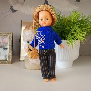 May include: A vintage doll dressed in a blue sweater with white accents, striped pants, and a straw hat. The doll holds a small basket and a wooden stick, suggesting a fishing theme. The doll is standing on a white surface.