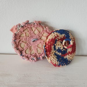 May include: Two handmade crochet items. One is pink with a floral design and a small pink ribbon. The other is a circular design with red, blue, and cream colors, with a small ring in the center.