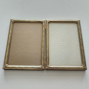 May include: A gold-colored, hinged picture frame with two panels. Each panel has a rectangular opening and a decorative border. The frame is designed to hold two photos or images. The frame is on a white surface.