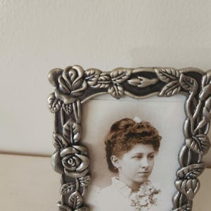 May include: A silver-colored picture frame with a portrait of a woman. The frame is decorated with roses and leaves. The portrait is in black and white.