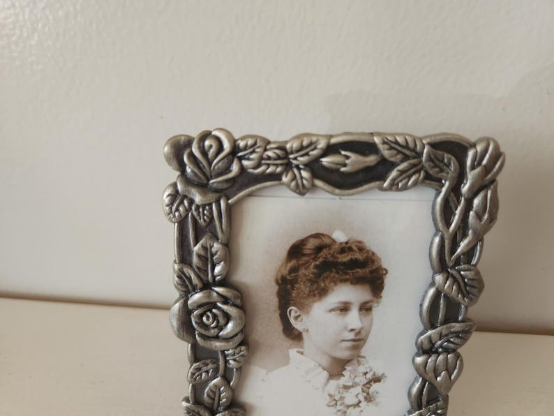 May include: A silver-colored picture frame with a portrait of a woman. The frame is decorated with roses and leaves. The portrait is in black and white.