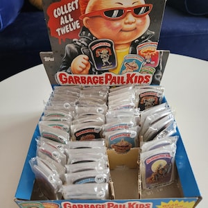 May include: A display box of Garbage Pail Kids buttons. The box features a cartoon image of a character with red hair and sunglasses. The buttons are individually packaged and feature various Garbage Pail Kids characters.