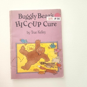 May include: A pink book with the title "Buggly Bear's HICCUPS Cure" by True Kelley. The book cover features a brown bear with a red bird, a blue bird, a rabbit, a raccoon, a fox, and a squirrel.
