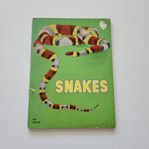 May include: A vintage paperback book with a bright green cover featuring a coiled snake with red, yellow, and black bands. The word SNAKES is printed in large yellow letters in the center. The bottom left corner displays 45 cents and TW176.