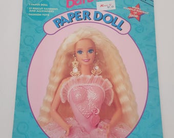 May include: A vintage Barbie Paper Doll Deluxe Edition book. The cover is teal with the Barbie logo in pink. The image features a Barbie doll in a pink dress. The book includes a paper doll, precut fashions, accessories, and a fashion tote.
