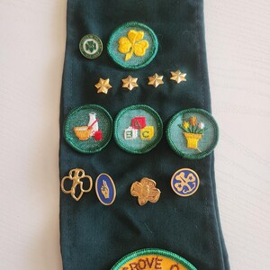 May include: A dark green sash with various embroidered patches and pins. Patches include a yellow shamrock, stars, and images of a baseball, ABC blocks, and flowers. Pins feature a gold trefoil and other designs. A large patch at the bottom reads "Yellow Grove Camporee."