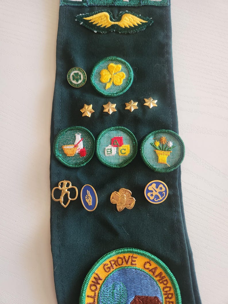 May include: A dark green sash with various embroidered patches and pins. Patches include a yellow shamrock, stars, and images of a baseball, ABC blocks, and flowers. Pins feature a gold trefoil and other designs. A large patch at the bottom reads "Yellow Grove Camporee."