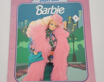 May include: A vintage Barbie deluxe paper doll book. The cover shows a Barbie doll in a pink fur coat and hat, with a teal purse. The background depicts a city skyline. The text reads "MORE FASHIONS!" and "more precut clothes fashions to finish storage envelope."