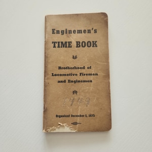 May include: A vintage, rectangular "Enginemen's TIME BOOK" with text in black. The cover is a faded brown with the text "Brotherhood of Locomotive Firemen and Enginemen" and the number "1949". The book is slightly worn.