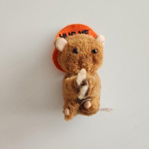 May include: A small, brown teddy bear figurine with an orange hat. The hat has the words "HUG ME" in black letters. The bear is holding its arms out as if to hug someone. The bear has small black eyes and pink ears.