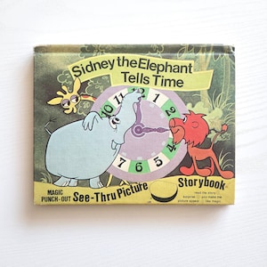 May include: A vintage children's book titled "Sidney the Elephant Tells Time." The cover features illustrations of an elephant, lion, and giraffe around a clock face. The book has a "See-Thru Picture" feature.