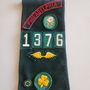 May include: A dark green sash with embroidered patches. The patches include the word "PHILADELPHIA" in red, a white flower, the number "1376", gold wings, a shamrock, a Girl Scout pin, and gold stars. The sash is likely for a Girl Scout.