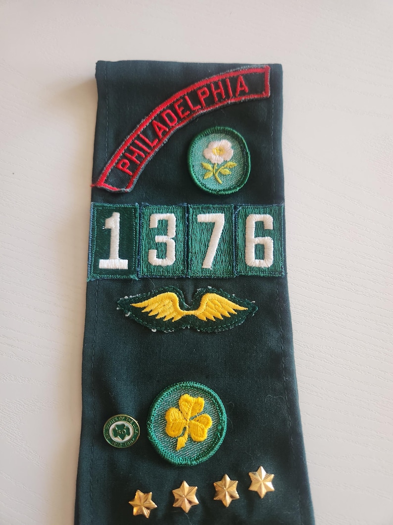 May include: A dark green sash with embroidered patches. The patches include the word "PHILADELPHIA" in red, a white flower, the number "1376", gold wings, a shamrock, a Girl Scout pin, and gold stars. The sash is likely for a Girl Scout.