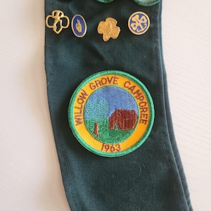 May include: A dark green sash displaying several Girl Scout badges and pins. The badges feature images of a beaker, ABC blocks, and flowers. Gold-colored pins and a round patch with the text "Willow Grove Camporee 1963" are also visible.