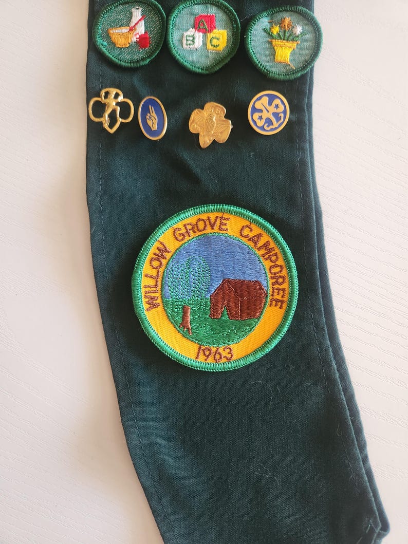 May include: A dark green sash displaying several Girl Scout badges and pins. The badges feature images of a beaker, ABC blocks, and flowers. Gold-colored pins and a round patch with the text "Willow Grove Camporee 1963" are also visible.